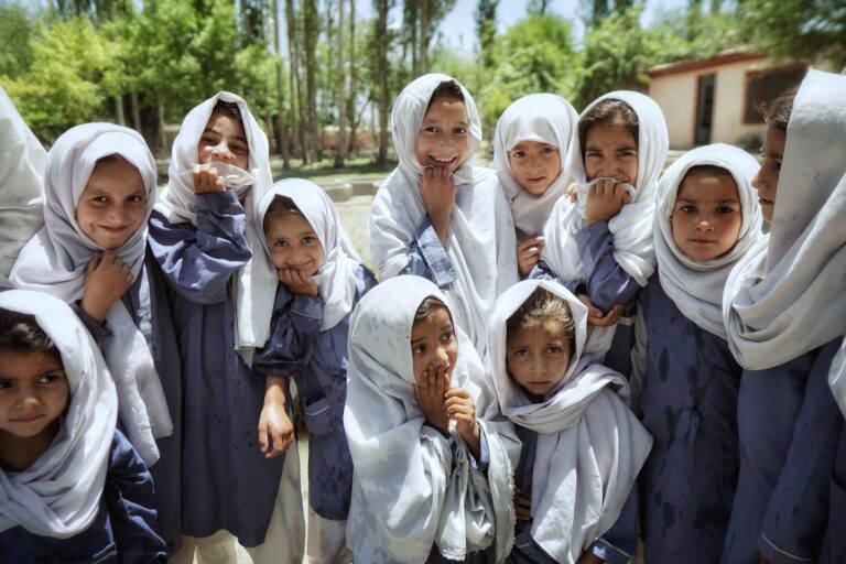 A cheerful group of children wearing traditional clothing and headscarves, smiling in the sun.