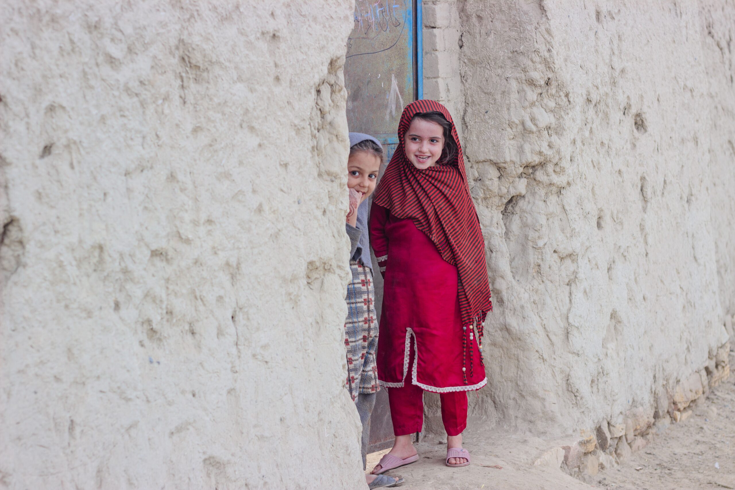 Two girls in colorful traditional dresses smiling while standing beside a textured stone wall.