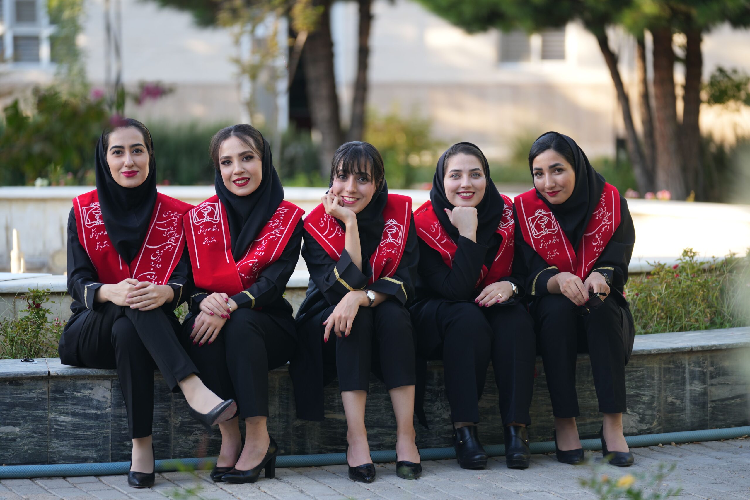 Five Iranian women in graduation attire, smiling outdoors, celebrate their achievement together.